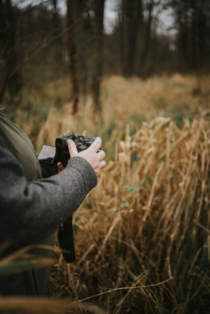 Zoey Elinor holder kameraet og ser motivet an, omgivet af høje siv. Brandingbilleder til hjemmeside i Jylland.