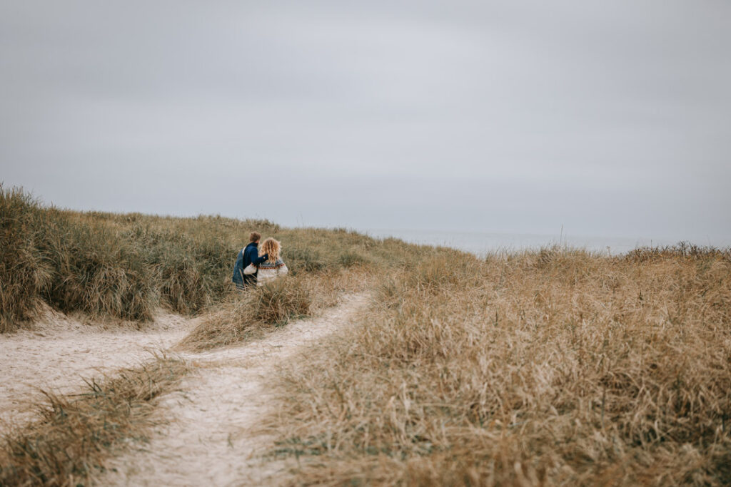 Par går hånd i hånd gennem marehalm ved stranden – parfotografering i Struer.