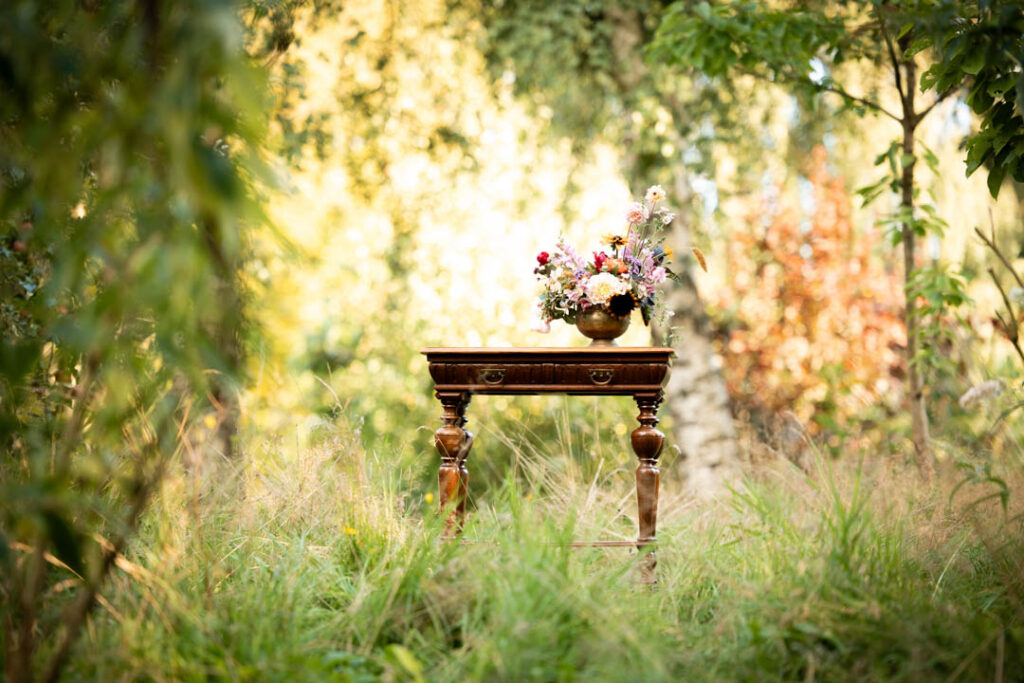 Eventfotograf fotograferer et lille antikt bord i skovlys med blomster i baggrunden.
