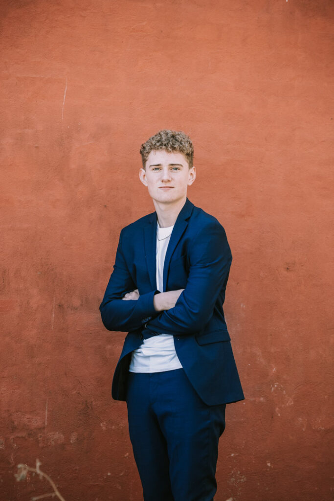 Halvfigurportræt af konfirmand i blå blazer foran varm, rustfarvet væg.