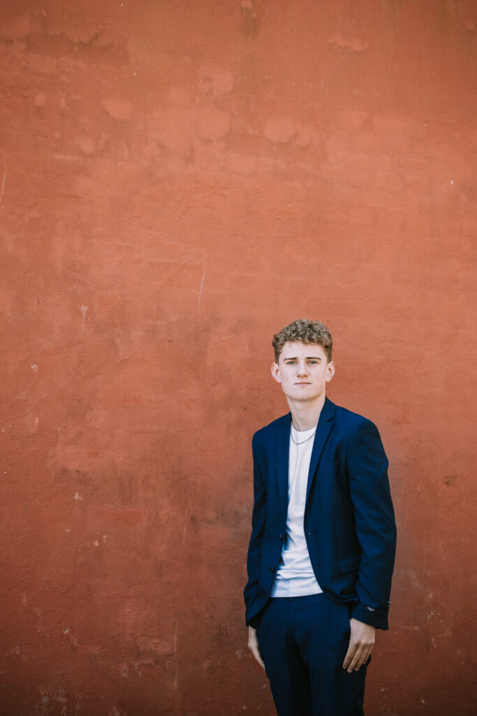 Konfirmand står alene foran rustfarvet baggrund og ser direkte mod kameraet. Konfirmationsfotograf Zoey Elinor