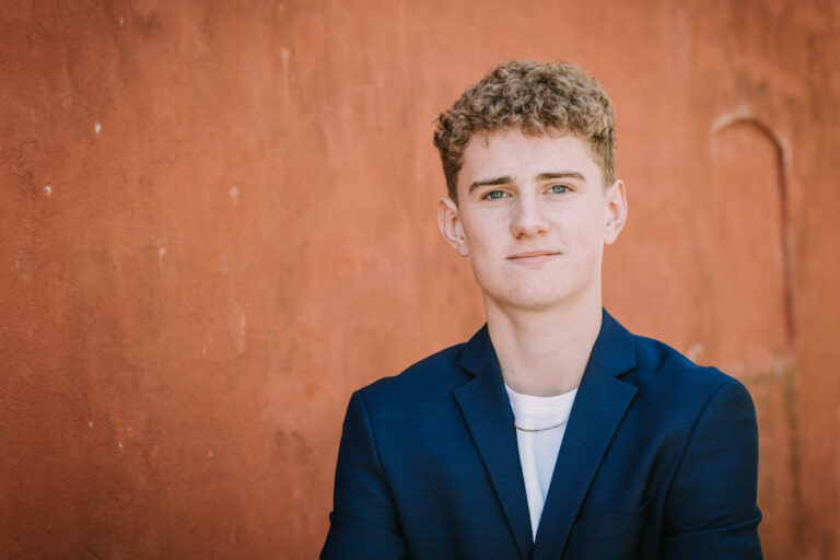 Portræt af konfirmand i blå blazer foran rustfarvet væg med afslappet udtryk.