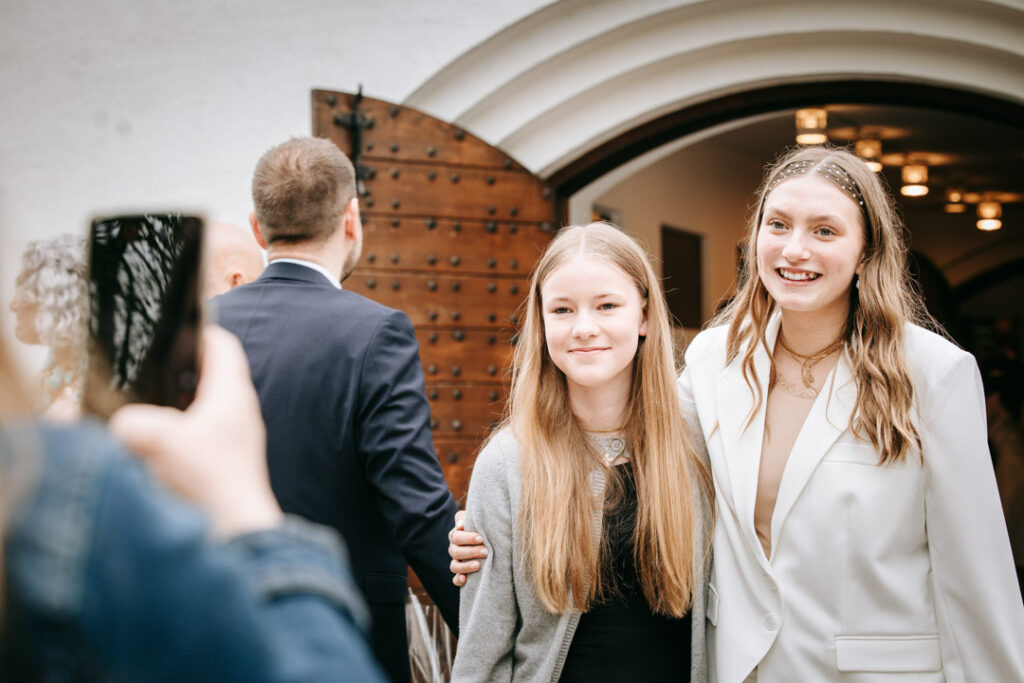 Konfirmand står sammen med veninder udenfor kirken og smiler, fanget i naturlige konfirmationsbilleder.