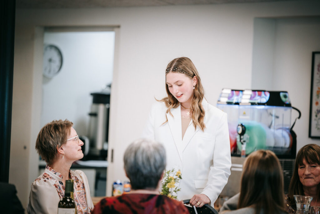 Konfirmand står og taler med gæster indendørs iført konfirmationstøj, fanget i et naturligt øjeblik under tale.