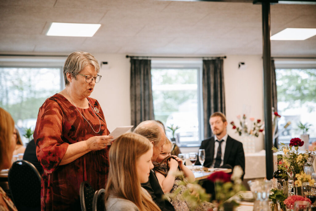 Gæster samles omkring bordet indendørs, mens konfirmationsfotograf dokumenterer stemningen og samværet.